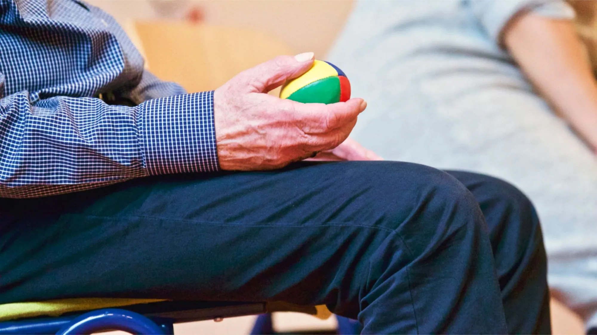 Ältere Hand hält einen bunten Ball auf dem Knie, kariertes Hemd